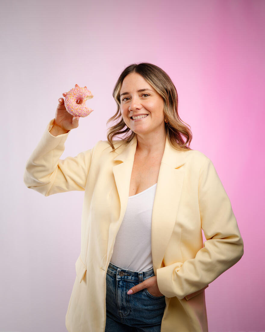 Océane en blazer jaune sur fond rose, un donut à la main